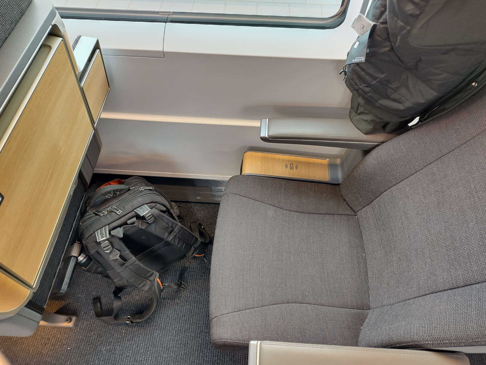 A picture of a first class seat on a Deutsche Bahn ICE train. It's in the companies new interior design. Instead of the previous blue leather color scheme, the new interior has the seat made of fabric of the same kind you can find on premium office chairs. The arm rests are still made of leather. Both the seat and the armrests are in gray instead of the traditional blue Deutsche Bahn has been using for its interiors for a long time. The seat pictured here is a single-row seat. In first class on Deutsche Bahn trains, each row has only three seats, in a 2-1 configuration. Between the seat and the window is a small-ish placement area held in light wood, with the universal contactless charging symbol on it. The appliances in front of the seat, a table and a cup holder, are both folded in this picture. Their backsides are held in the same light wood as the wireless charging area. Overall, the seat area makes a very premium impression on me.