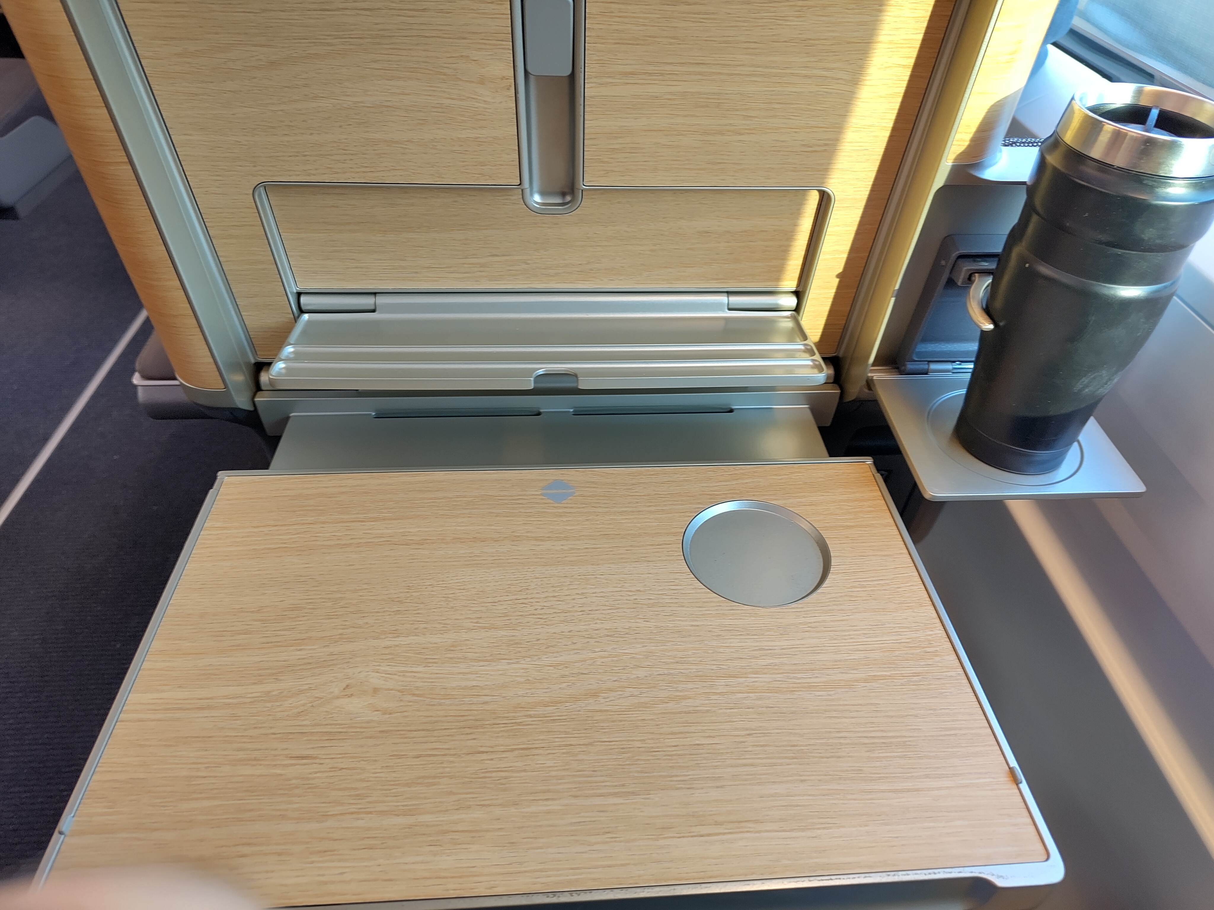 Another picture, this time made from the perspective of somebody sitting in the seat. The folding table and drink holder are now extended. The table's surface is made from the same wood as the backside, and when folded out, the area previously covered by the table is held in the same wood. The top of the cup holder is made of metal. The table has another slight indent for another cup. Above the table is a smaller appliance folding out, a lot shorter than the table and with some groves in it, made for holding a phone or a tablet. At the top of the picture is a metal hook which can be used together with the phone holder to keep the phone/tablet in place.