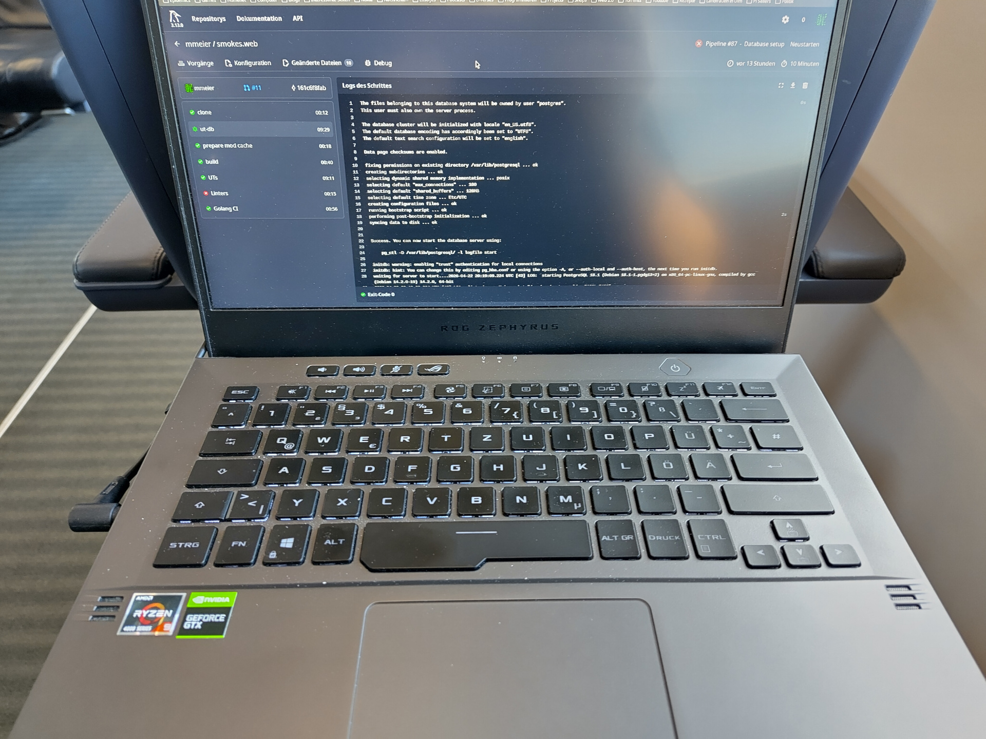 A picture of a gamer-y laptop sitting on the tray table of a first class seat in a Deutsche Bahn ICE train. The screen shows the Woodpecker CI web UI for a pipeline run in the Smoekweb project. It shows multiple jobs, some successful, others not. The most interesting one is called 'ut-db'. It is not a normal job, but a service. Its logs are currently open, and it shows the startup of a Postgres database instance.
