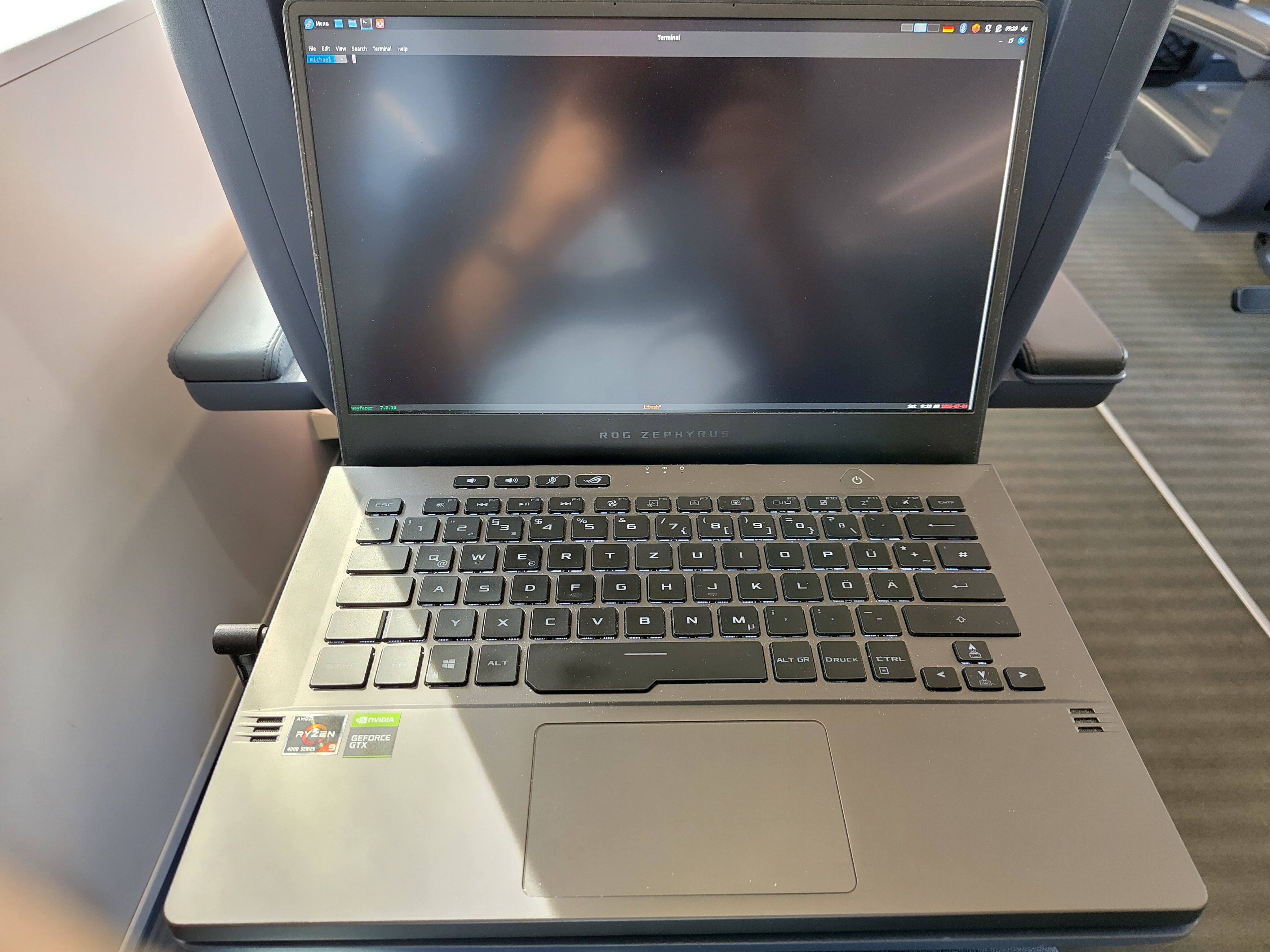 A picture of a gamery Asus laptop. The machine is resting on the tray table in front of a Deutsche Bahn ICE seat. The color scheme is still the older dark blue leather. The screen shows an open terminal, with only the prompt with the username 'michael' visible at the time. Desktop environment is Sway.