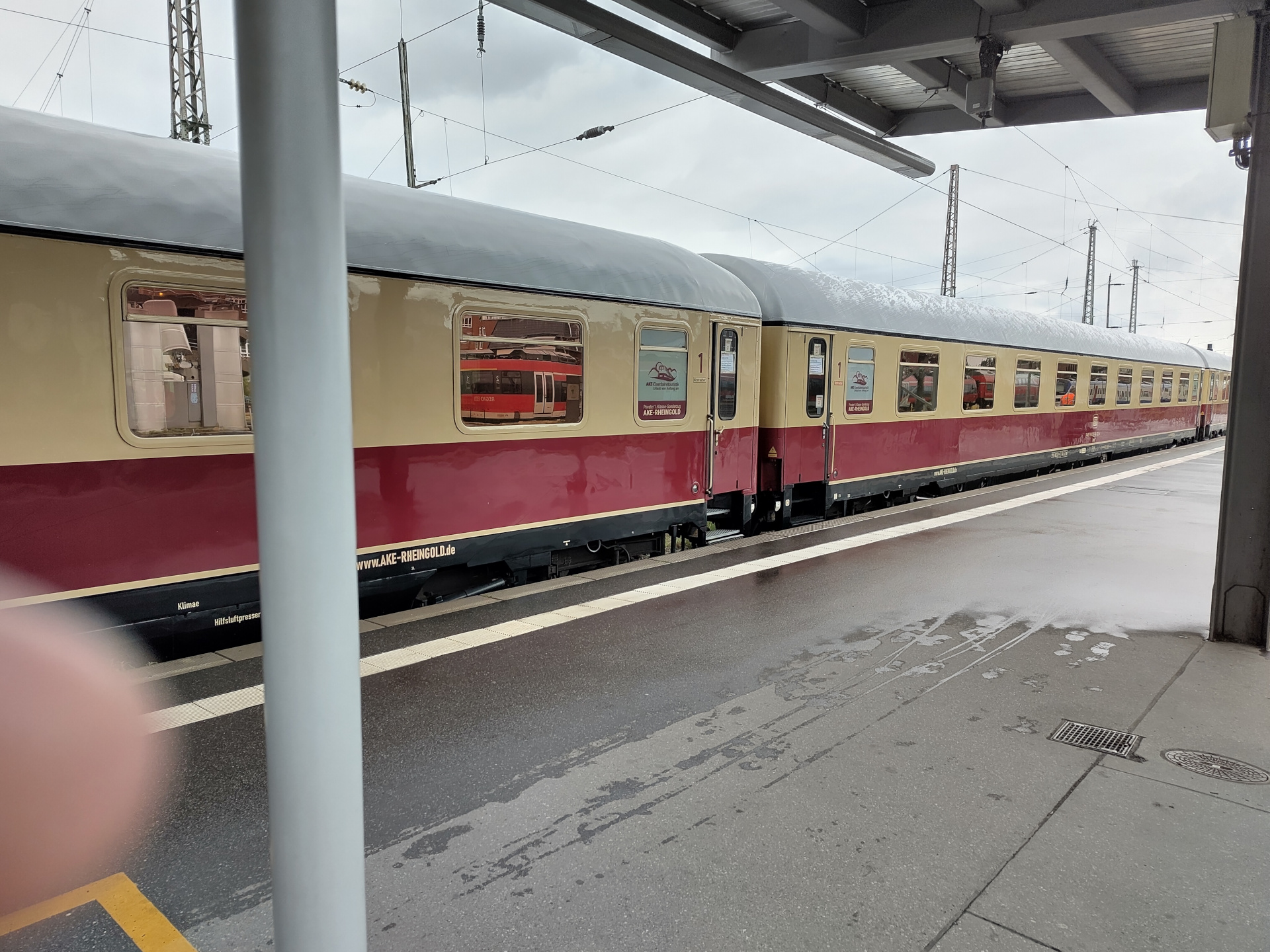 A picture of a few train carriages standing at a platform. The train is wearing a beige and wine red livery. A placard in the train's windows says, in German: 'AKE Train Tourism, vacation from the start', with 'Private 1st class special train AKE-RHEINGOLD'. The carriages are definitely of older vintage than Deutsche Bahn's normal stock, but still do look well cared for.