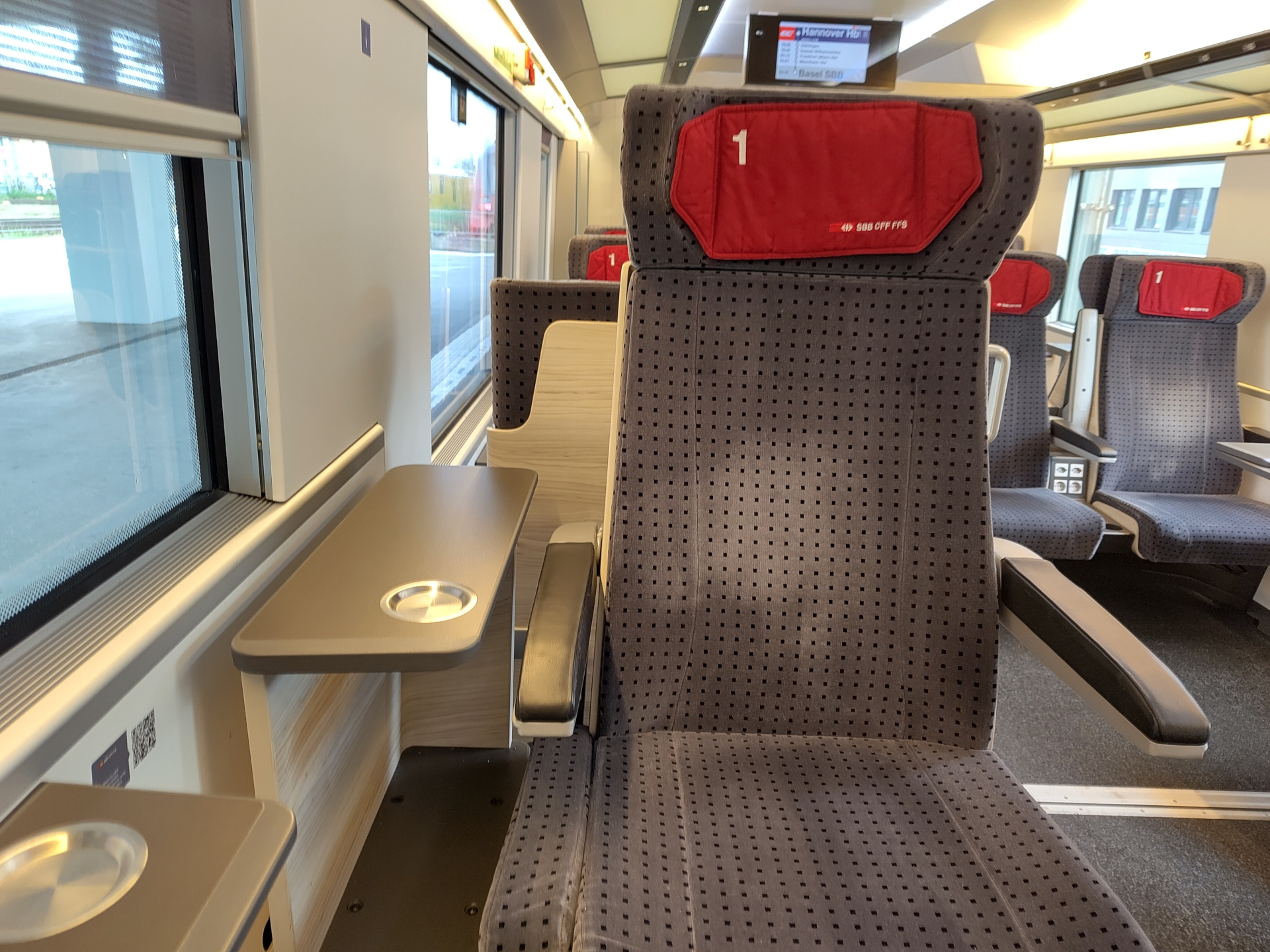 A picture of the interior of a train. In this case, not a Deutsche Bahn one. Instead, it's the interior of a SBB train. Instead of the leather seats customary in Deutsche Bahn's first class, these seats are made out in fabric. The color of the seats is gray, with black dots. The headrest has a red cushion with the SBB logo. To the right of the seat is a small table with a cup holder, but markedly, there is no table or tray in front of the seat.