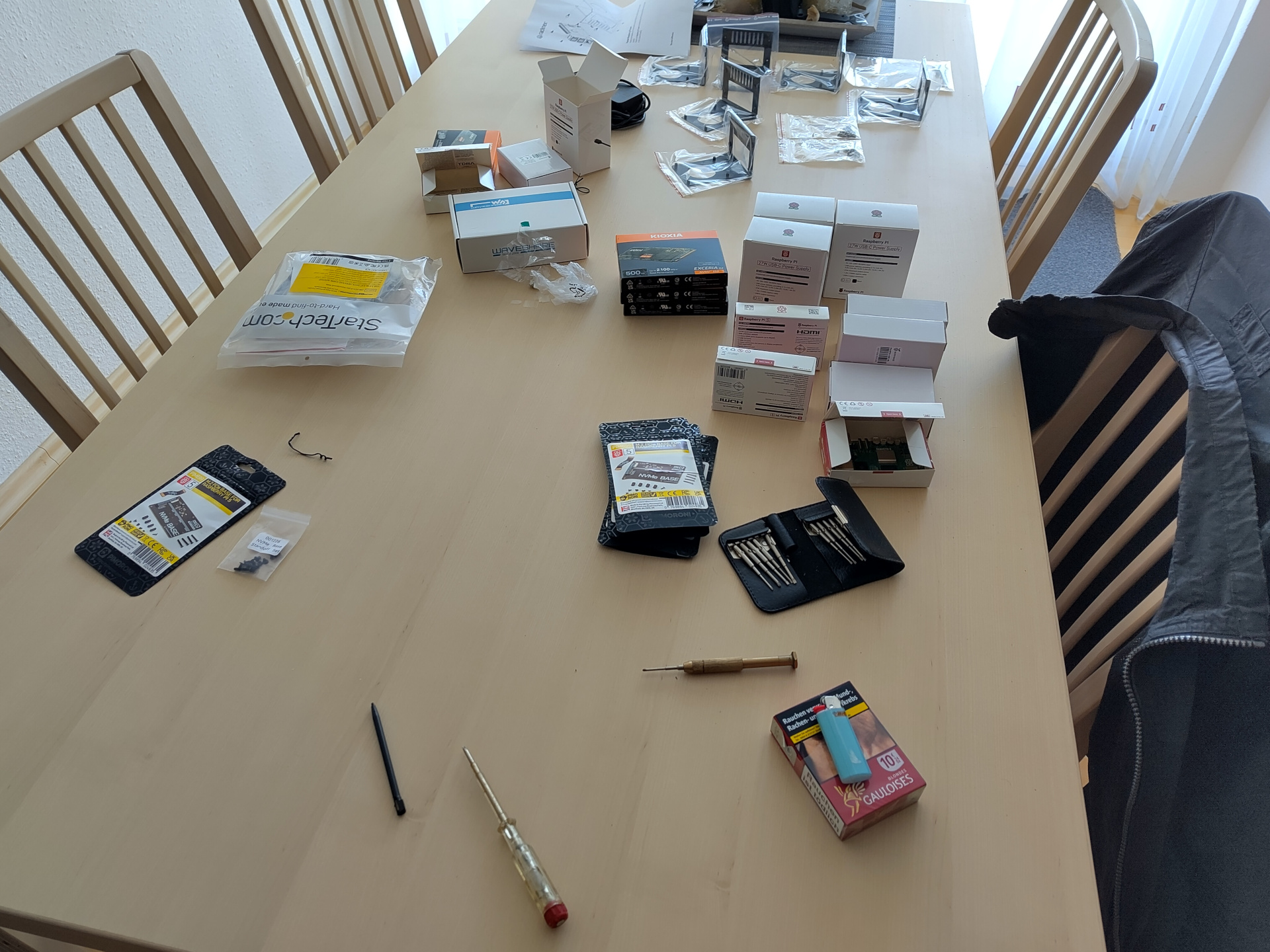 A picture of a table laden with Raspberry Pi components. There is three of most things. Three 500 GB Kioxia NVMe SSDs. Three Raspberry Pi 5 8Gb. Three official Pi 5 27W power supplies. Three Pimoroni NVMe baseplates. The one exception are the Racknex mounting plates. Of those there are six. Also visible in the foreground is a Hama precision screwdriver set.