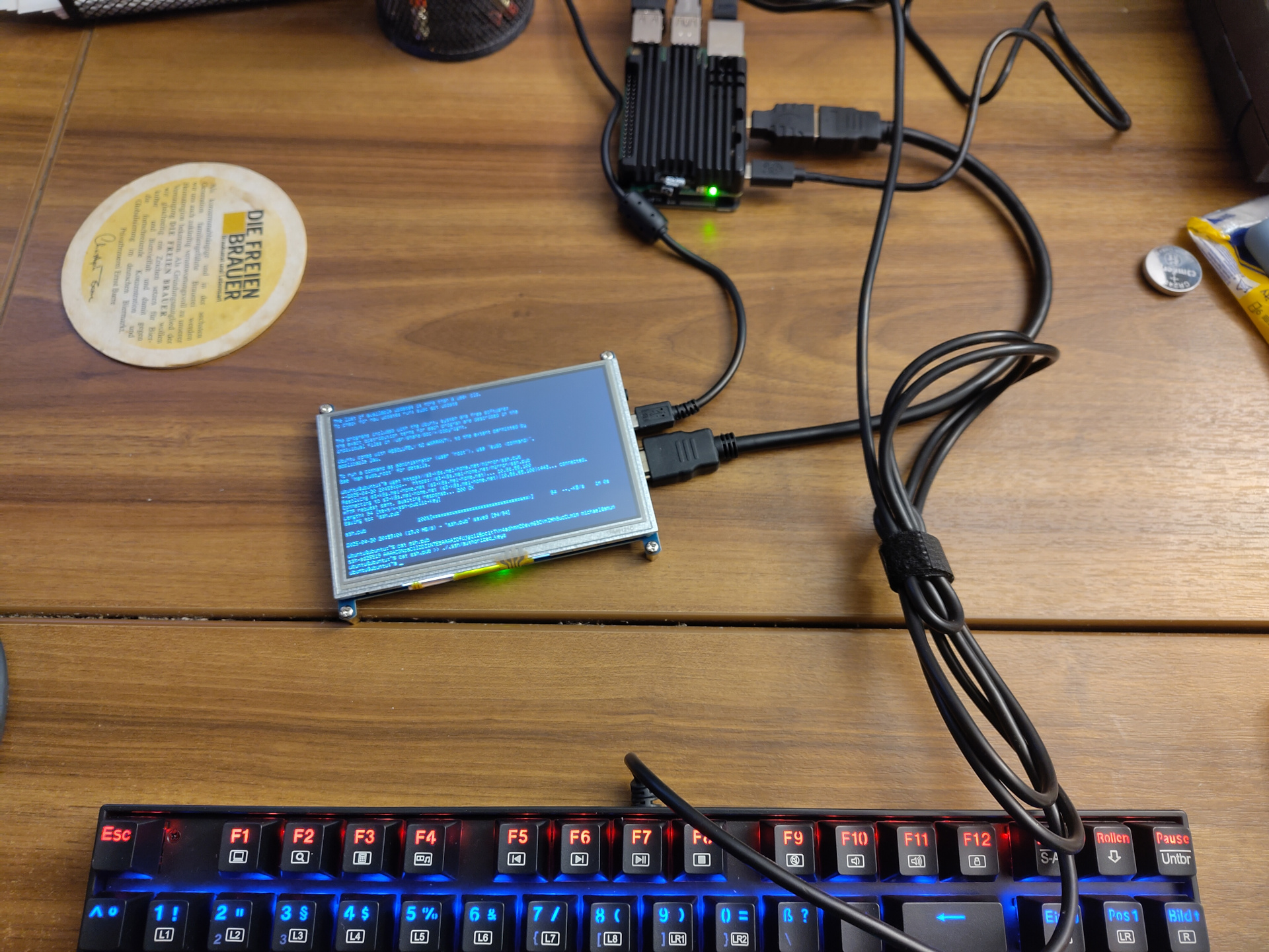Another picture of a Pi 5. The Pi itself looks similar to the other pictures. The important difference here is that it's sitting on a desk. It is connected to a relatively small TKL keyboard with a wonderful amount of rainbow puke going on. The center piece is a small 5 inch display. It is connected to both, a USB port and a HDMI port on the Pi. Squinting a bit, the text on the screen is legible, showing a terminal session with a download of an SSH public key and copying that key into the user's authorized_keys file.