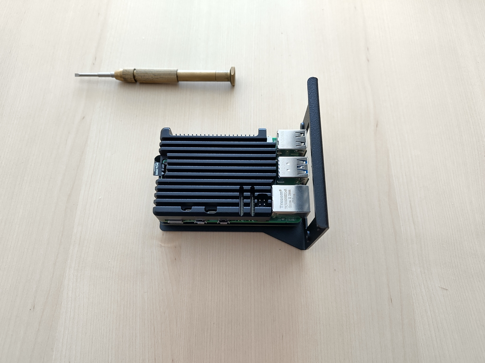 A picture of a Raspberry Pi. The Pi 5 itself is covered with a black with a black aluminum heat sink which is about as high as the front IO connectors and covers the entire board, with some cutouts for access to connectors. At the back, a PCIe cable is going from the Pi down to the NVMe base mounted below the Pi. The cables flimsiness and shortness screams 'I am a pain to handle'. The entire assembly is mounted onto a sturdy metal piece, with a front part angling up to about two Pis in height, with a large cutout for the Pi's IO in the front.