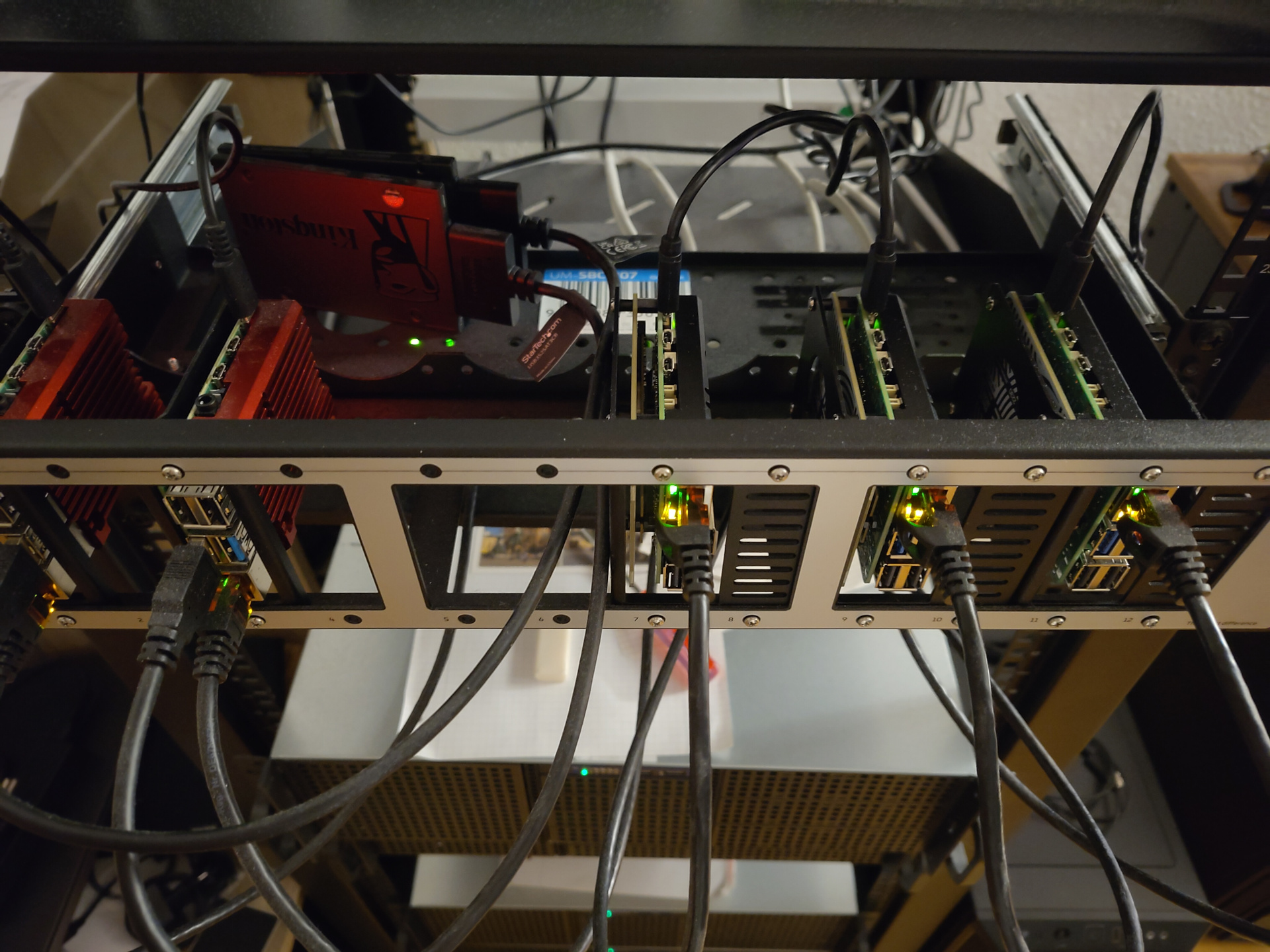 A picture of a Racknex Pi mount in a 19 inch rack. There are twelve slots to mount Raspberry Pis, with 8 currently occupied. On the very left are two Pi 4, each occupying one slot. They are each covered by a large read heat sink. Each one is connected to a SATA SSD via a USB-to-SATA adapter. The SSDs are mounted vertically behind the Pis. On the right side, six slots are occupied with three Pi 5. Each of them has a network cable plugged in. They are covered with a black heat sink. There is definitely not a single speck of dust visible in the entire picture. Not one. You definitely cannot see the outline of three more SATA SSDs vertically mounted until recently behind the three Pi 5.