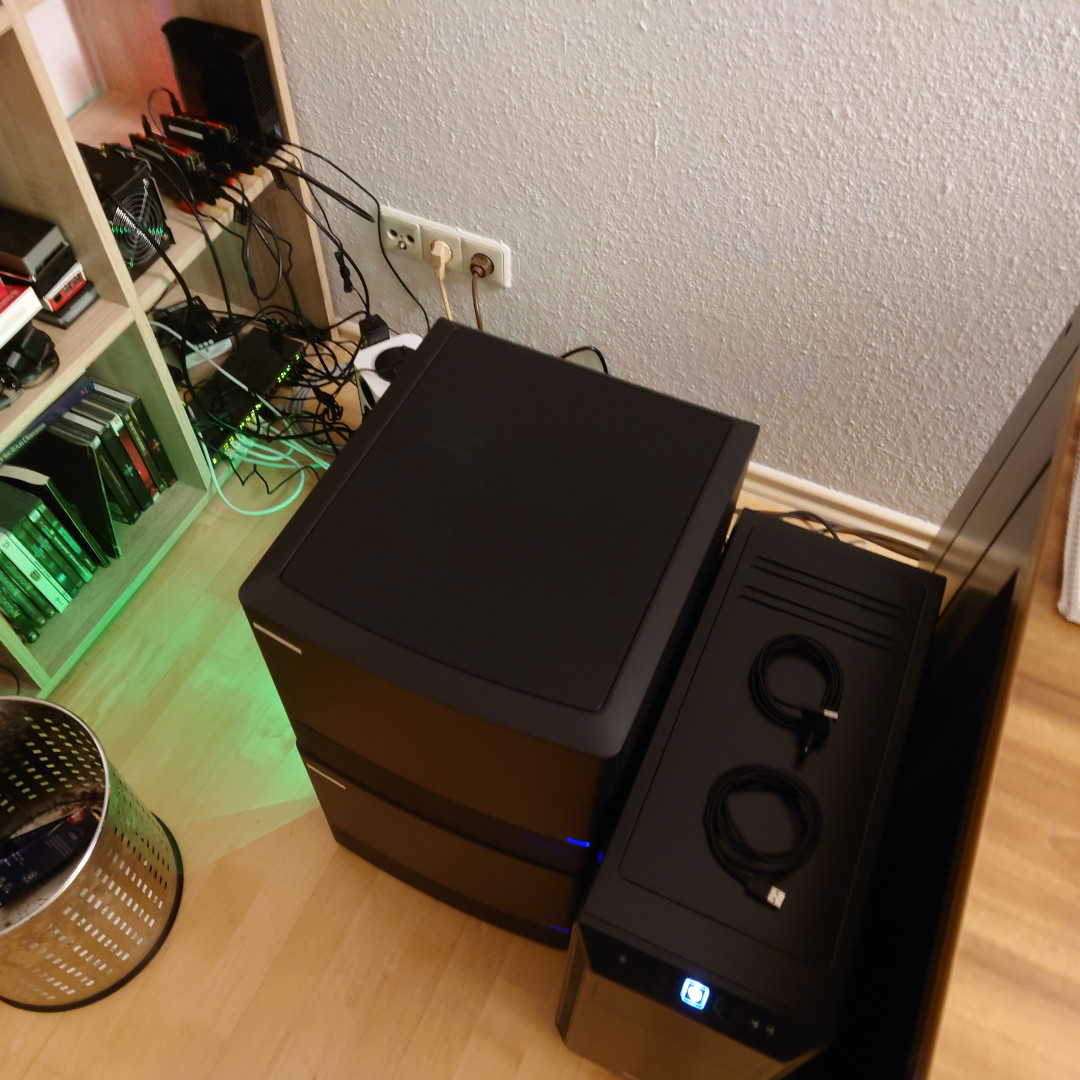 A picture of three computer cases, two of them stacked atop one another. To the right of them is another large big tower computer case. And to the right of it is part of a desk. To the left of the two stacked cases is a shelf. There are several SBCs and a network switch in it. On the ground between the stacked cases is a 12x power plug tower. Almost all outlets are used. Around all of it is a veritable jungle of cables.