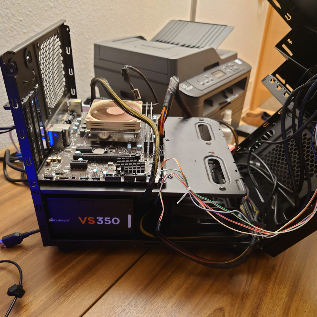 A picture of an open PC case. In it are two chambers. The lower one contains a power supply alongside a nest of cables. The model Corsair VS350 is noted on the side of the PSU. In the upper chamber is a brown mainboard, topped by a horizontal, low profile cooler in the proud brown and beige colors of Noctua.