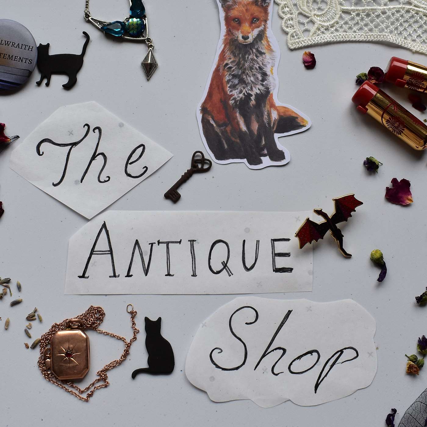 A large number of things on a white table. Several black cat stickers, a red fox sticker, a steel key, a metal figurine of a dragon, an amulet, a locket and a number of rose petals. All of these things are arranged around the name of the show, 'The Antique Shop'.