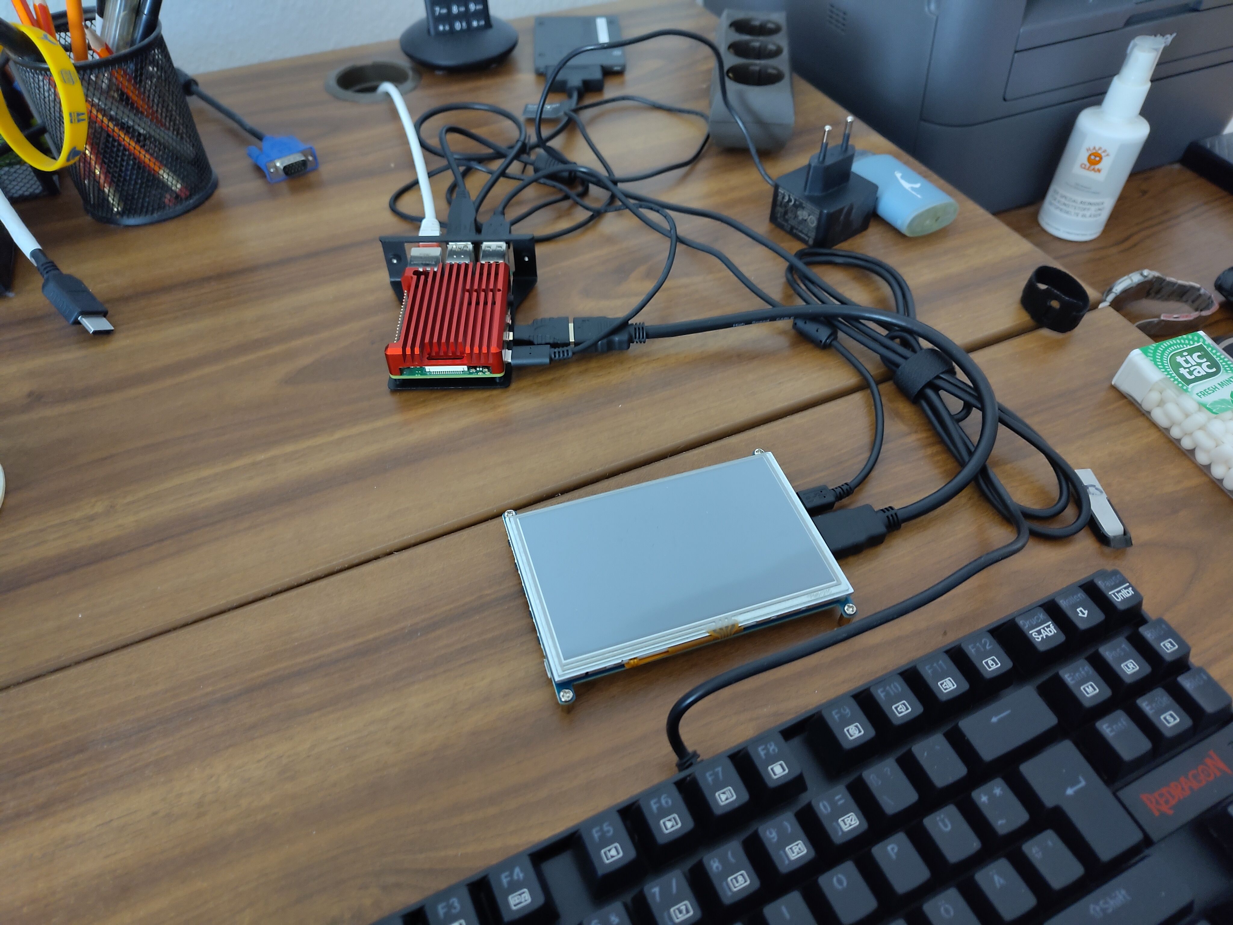 A picture of a desk with a Raspberry Pi 4 board and accessories. The Pi 4 is clad in a passive red heat sink and mounted on a right-angle piece of metal. It's connected to a small 7 inch screen with an HDMI and an USB cable. Furthermore, it's also connected to a keyboard and has a network cable plugged in. Finally, it's also connected to a 2.5 inch Kingston SATA SSD via a USB-to-SATA adapter.