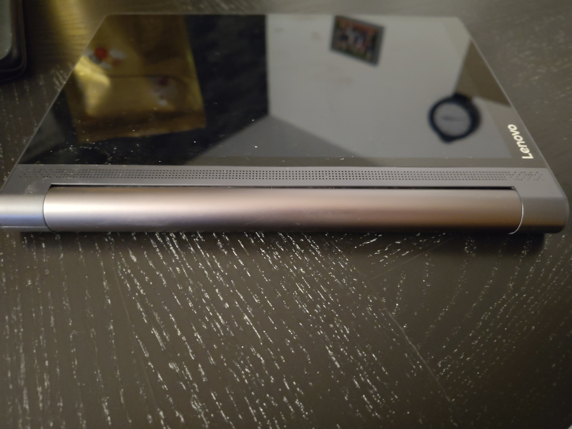 A picture of a Lenovo Yoga Tab sitting on a black table. It varies a bit from the typical 'black rectangle' aesthetic of Android tablets, in that it doesn't just consists of the screen. It also has a rather bulbous hinge+battery part on the long/left side, with a large speaker grill between that hinge and the screen. Due to that bulbous hinge, it doesn't lay flat on the table.