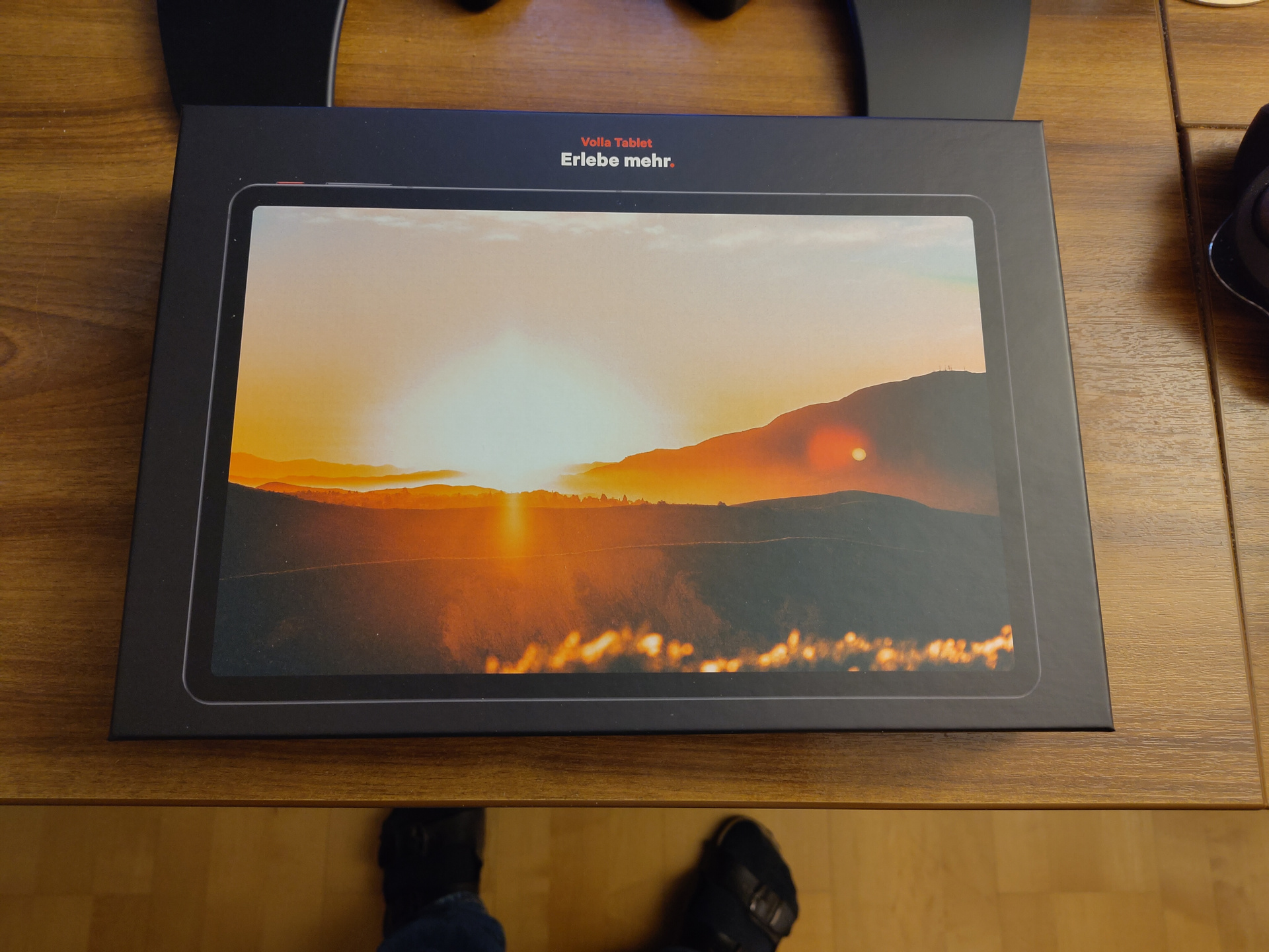 A picture of the Volla tablet package sitting on my desk. It is a black rectangle with a picture of the tablet on the front, with the slogan 'Volla Tablet' and 'Erlebe mehr' (meaning 'Experience more' in German) below that. The tablet itself is a very archetypical black rectangle. In the picture, it shows the dawn over a hilly and wooded area. In short, if you've ever seen an Android tablet, you know exactly what this one looks like.