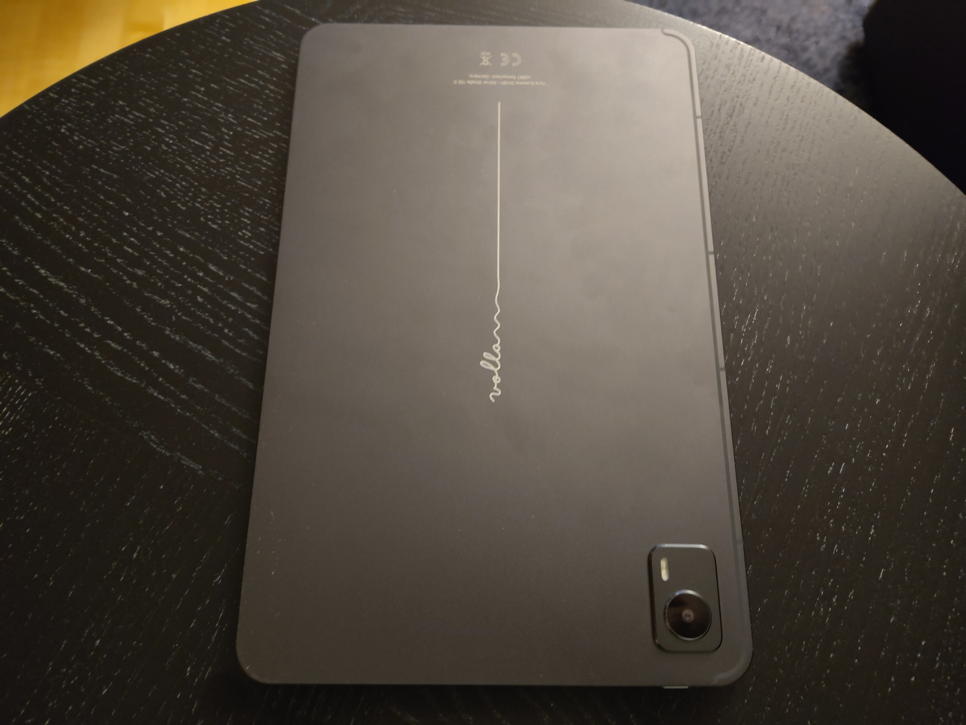 The same tablet on the same table, but now showing the back of it. The back is made of the typical light-gray metal, and it has a camera bump in the top right corner. The only distinguishing part of the design is the word 'Volla', written in cursive on the back, and after it a long line, as if somebody had handwritten the word and then just continued in a line down the center of the tablet.